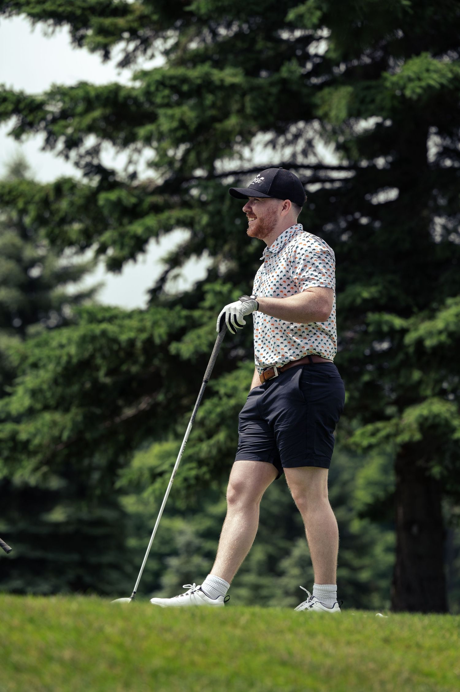 Golfer in checkered shirt and black shorts stands with golf club on grassy hill near evergreen trees.