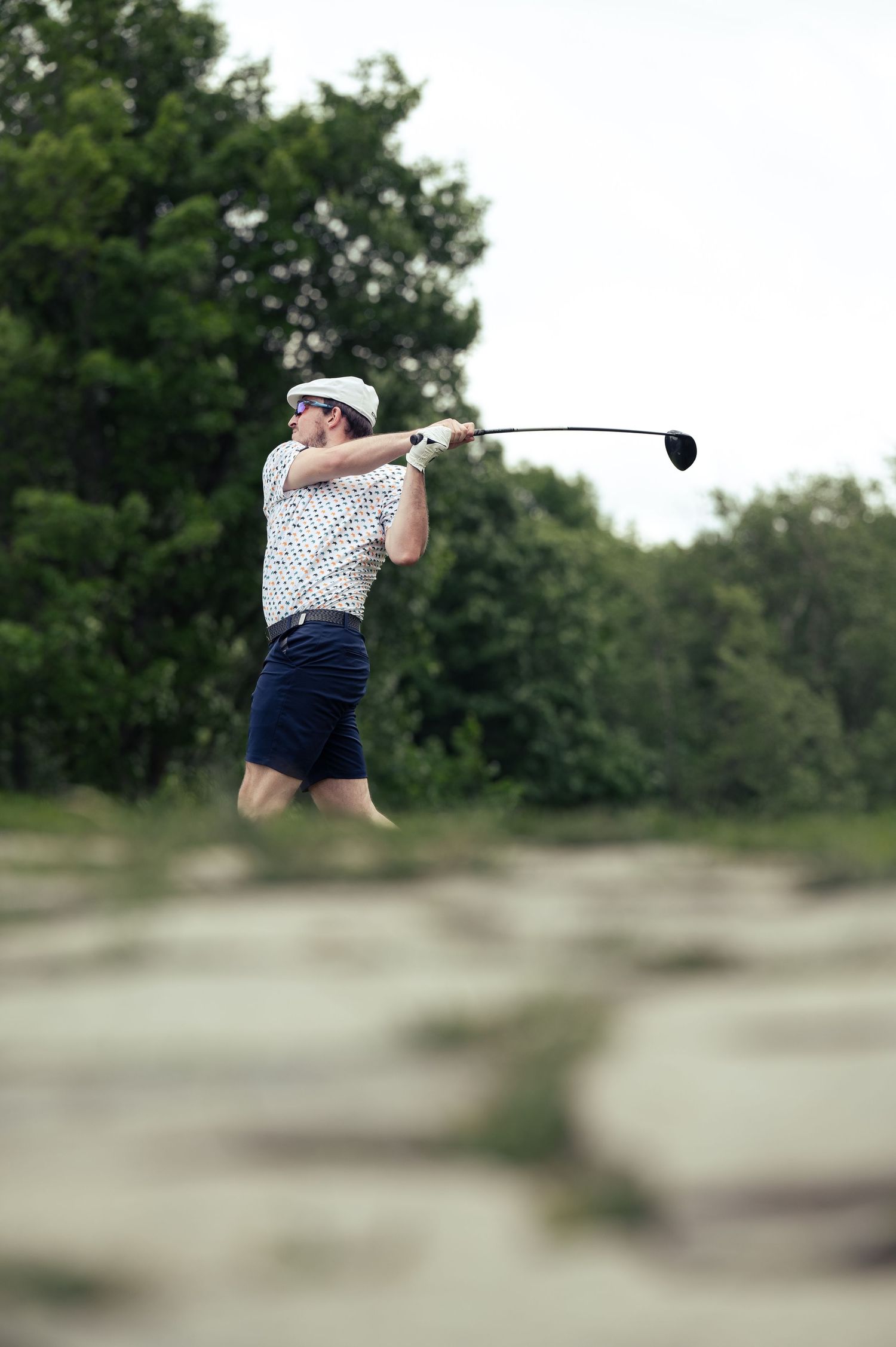 Golfer in vintage attire swings driver on golf course in front of tree-lined background.
