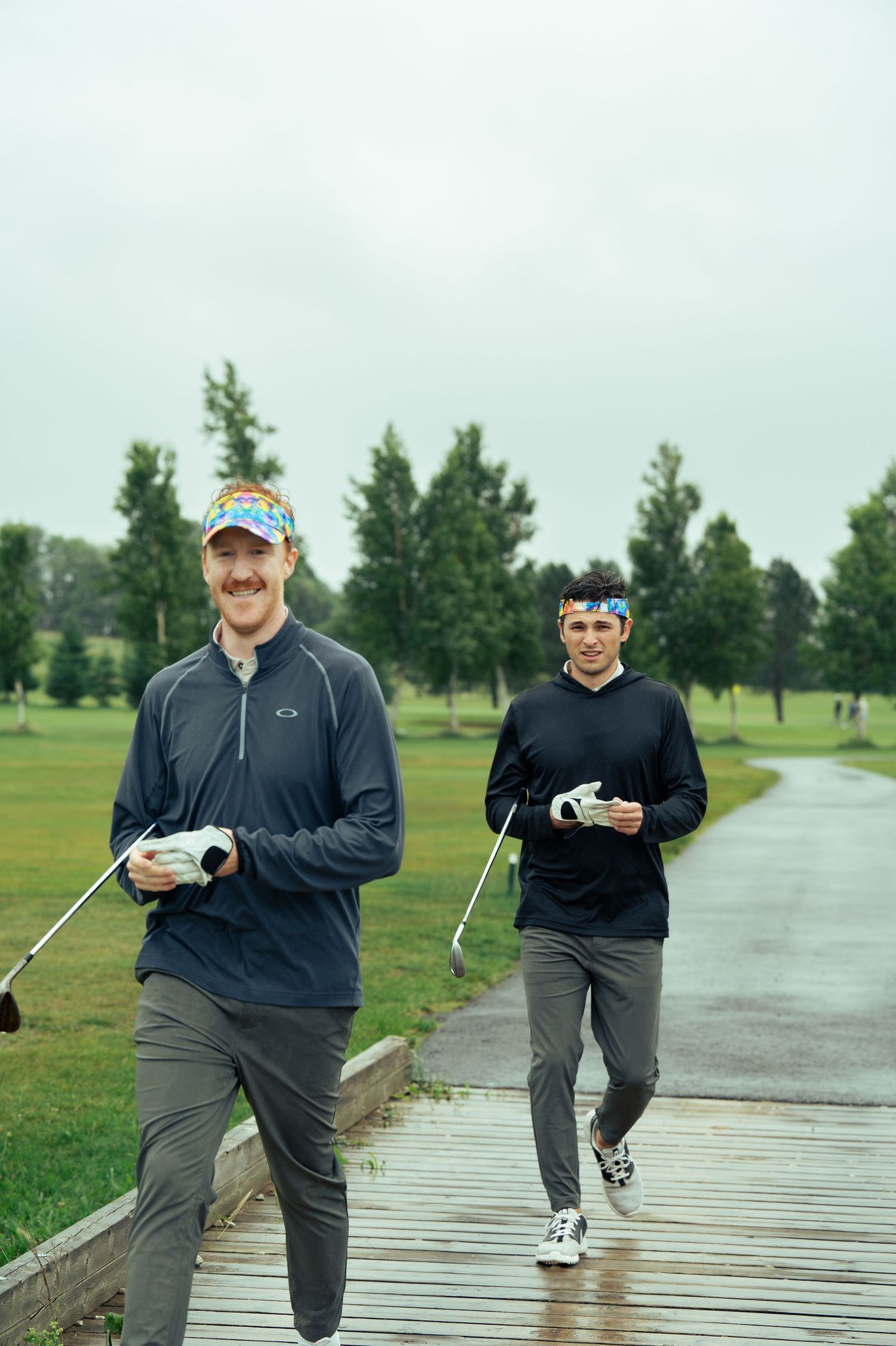 Two golfers walk along a wet cart path on a rainy day at a golf course surrounded by evergreen trees.