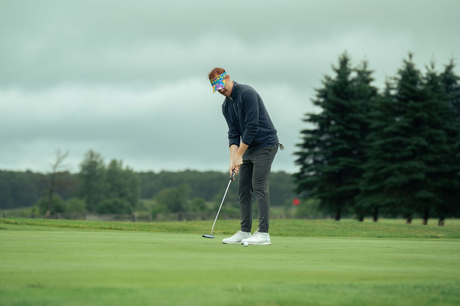 Golfer practicing putting on a cloudy day at a lush green golf course with evergreen trees in the background.