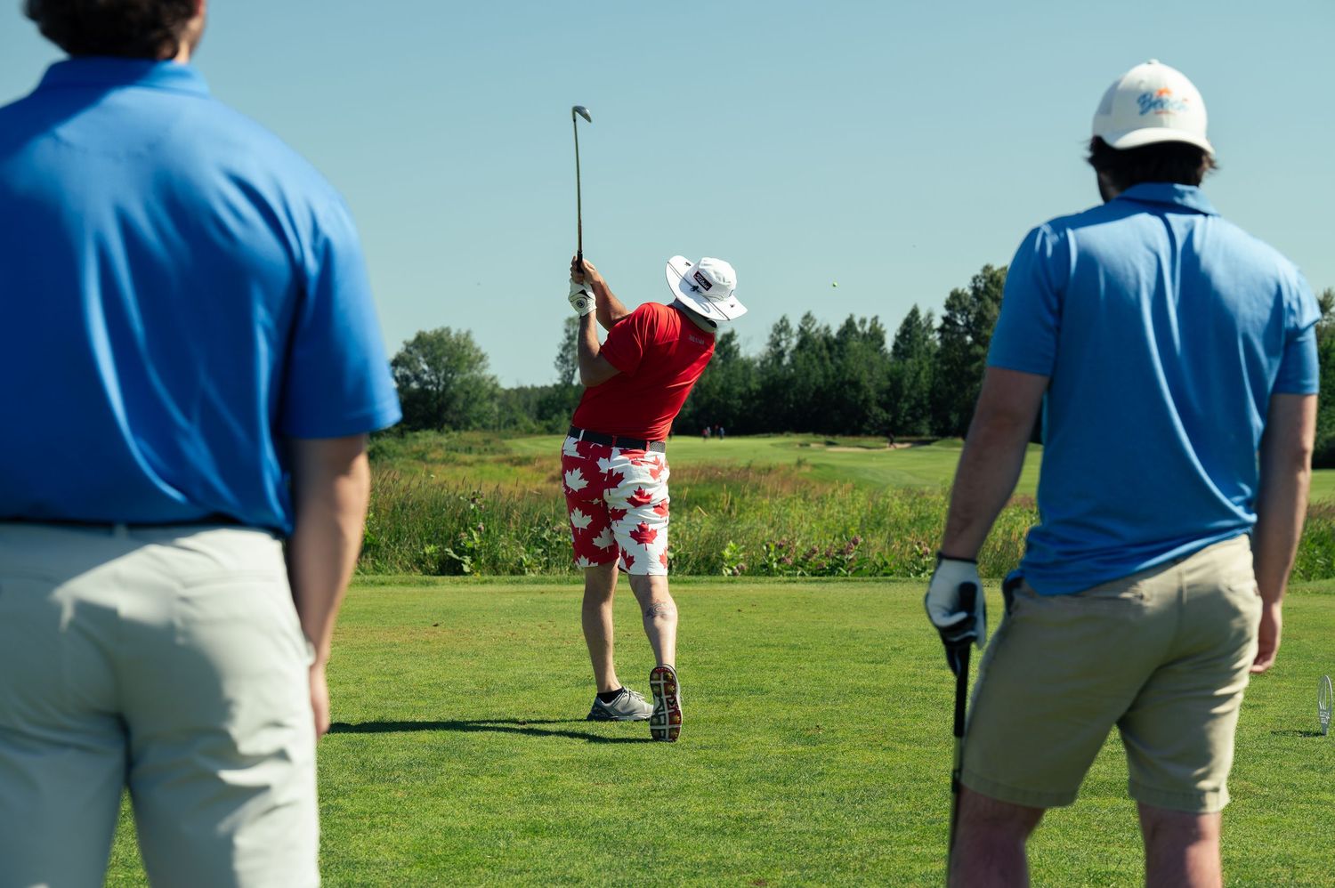 Golfers watching a player in red shirt taking a swing on a sunny day at the golf course.