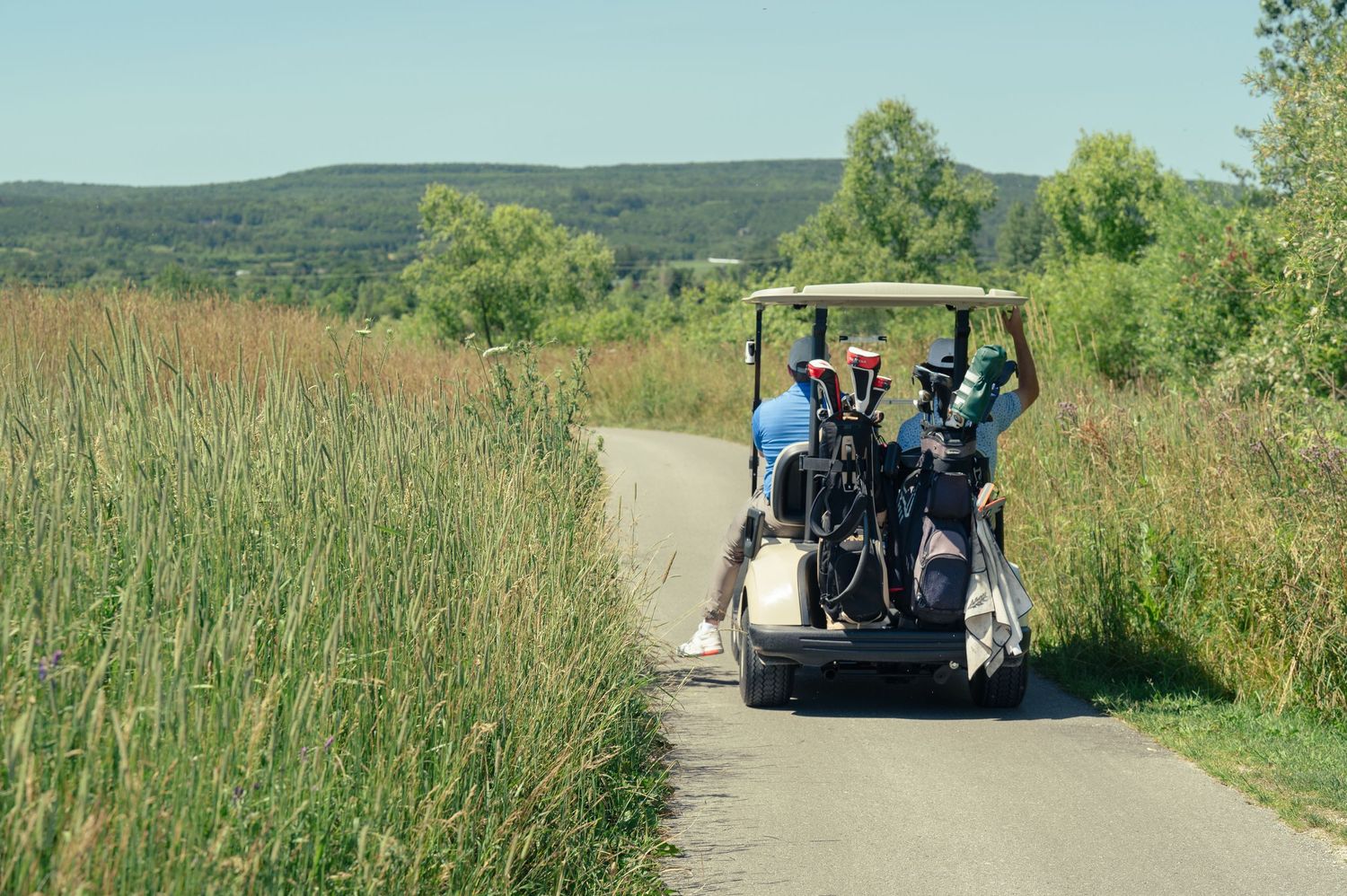 Golf cart carries passengers down a scenic path surrounded by tall grass and rolling hills on a sunny day.