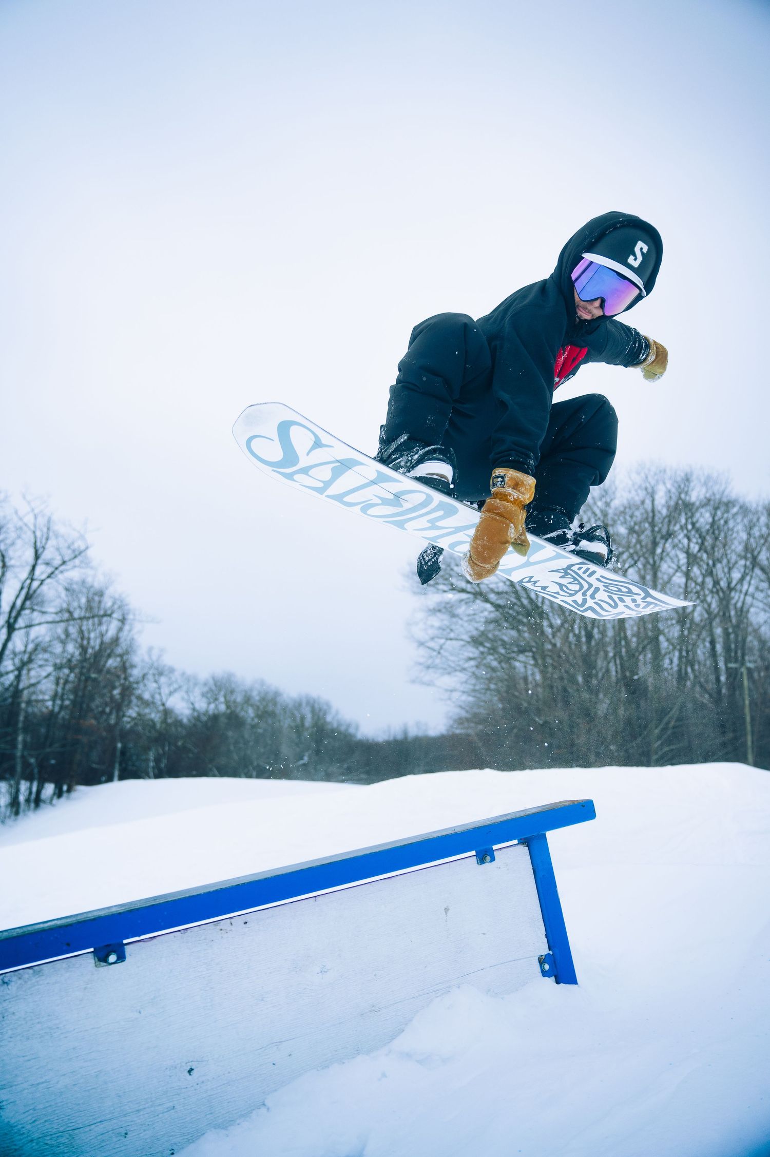 Snowboarder performs aerial trick sequence over blue terrain park rail in winter mountain setting.