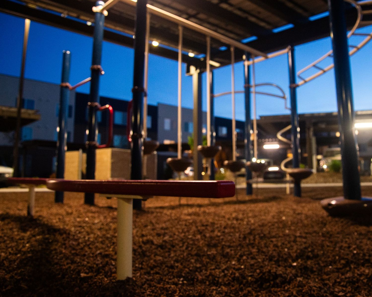 Colorful playground equipment at dusk with rubber mulch ground cover and modern building in background.