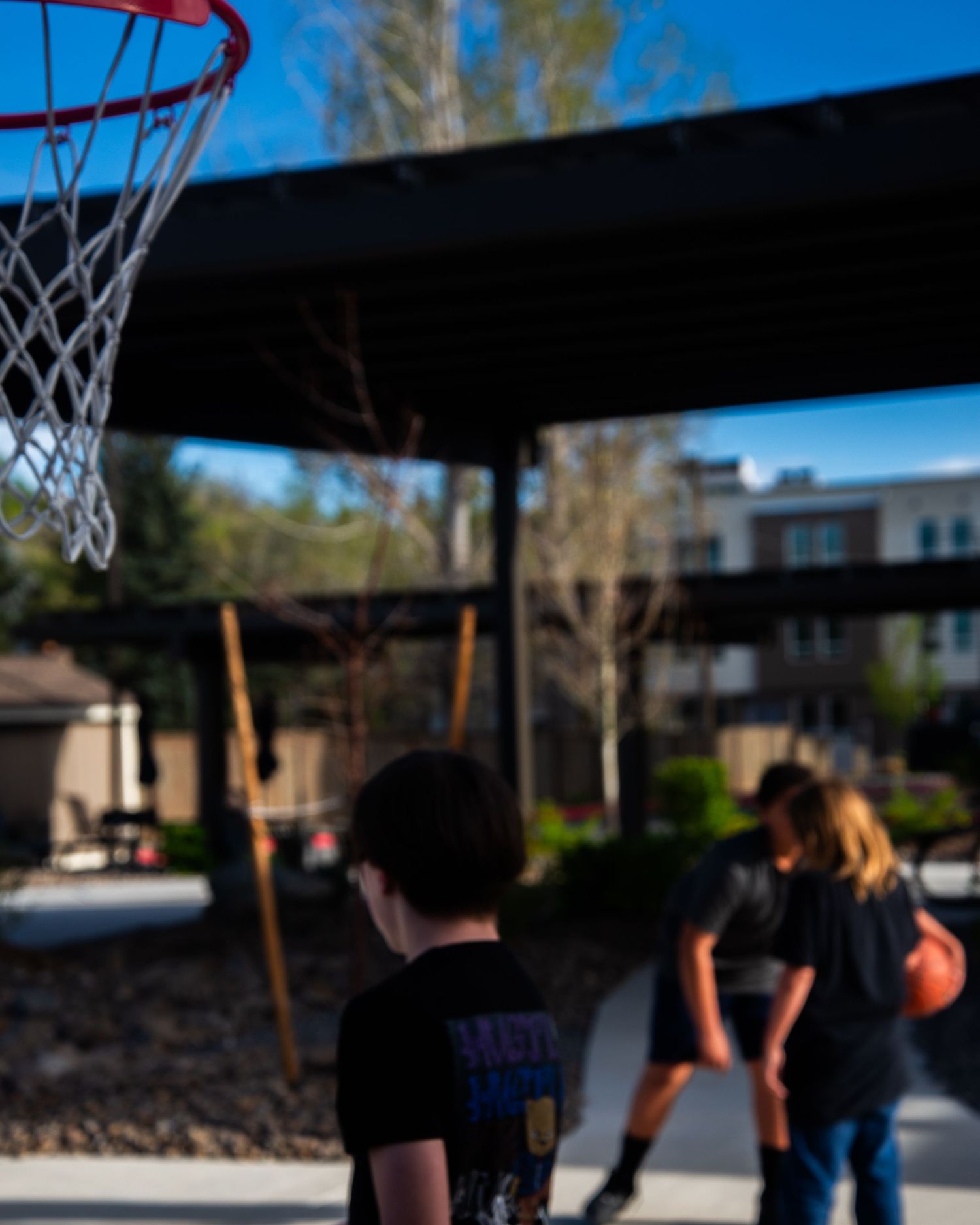 Two children playing basketball outdoors under a covered court surrounded by trees and residential buildings.