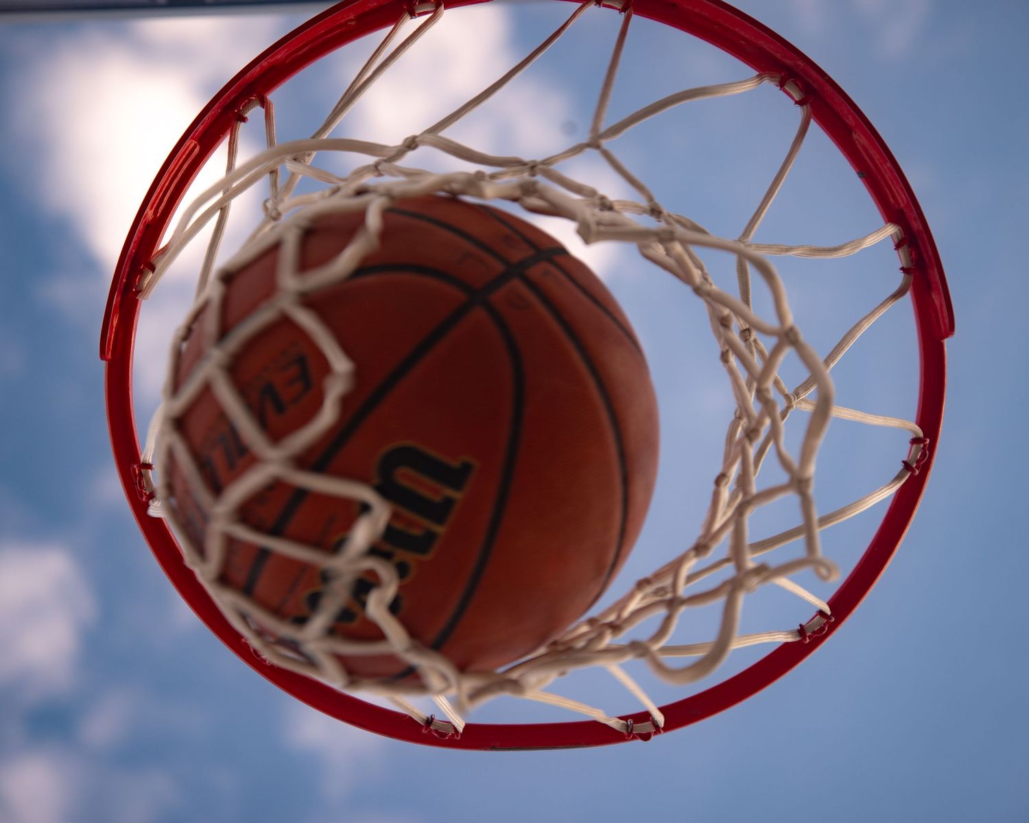 Close-up view looking up through a basketball hoop net as a Wilson basketball falls through against blue sky.