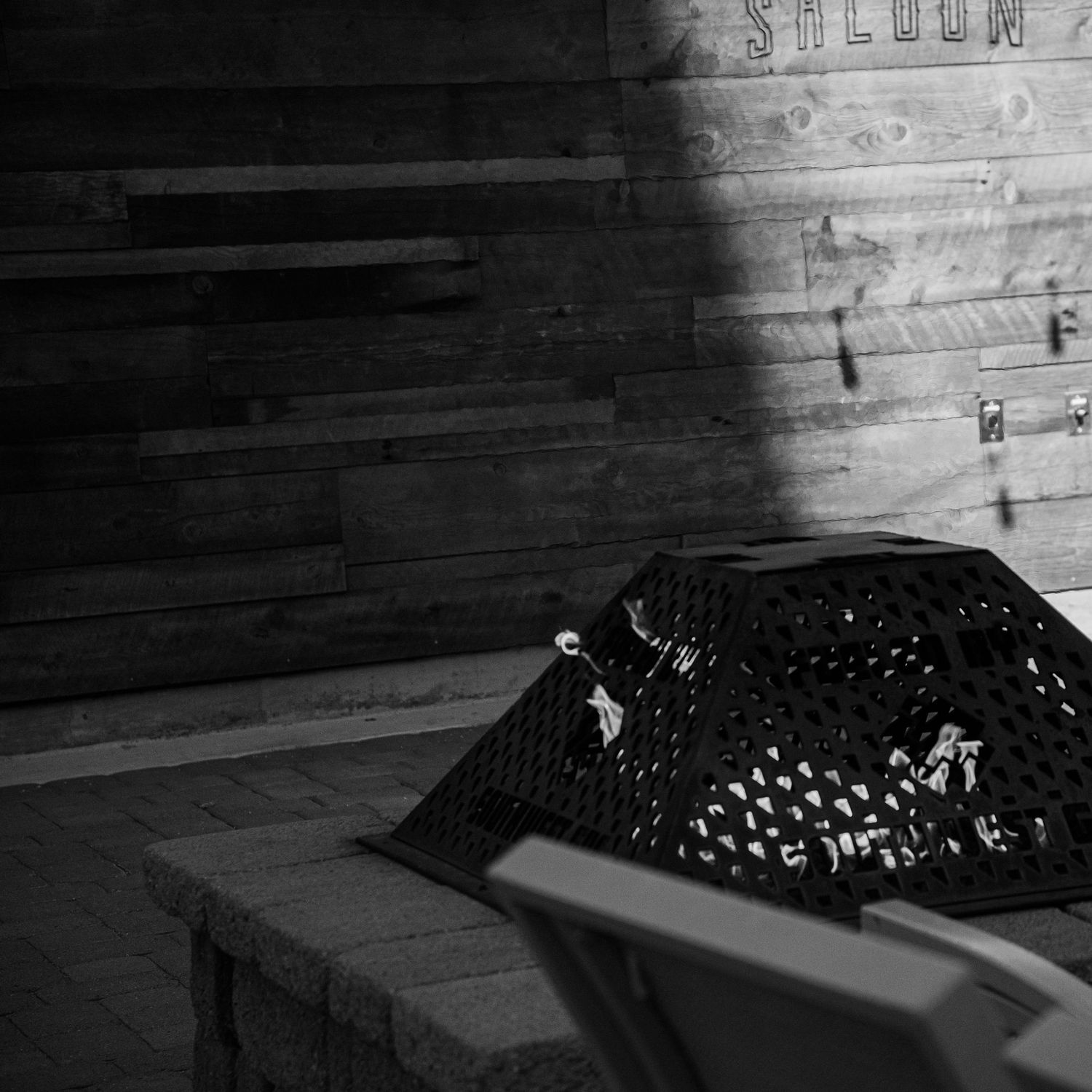 Black and white photo of a decorative metal fire pit cover on a brick patio near a rustic wooden saloon wall.