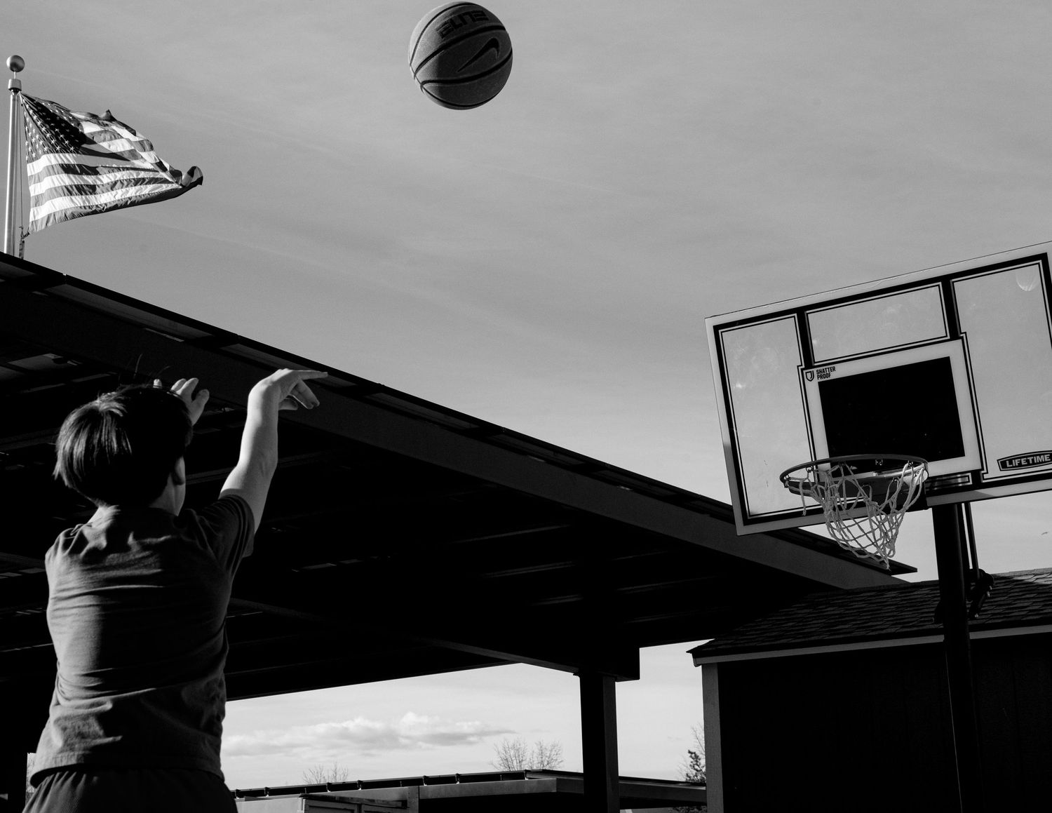 Boy shoots basketball toward hoop outdoors with American flag visible in black and white photo.