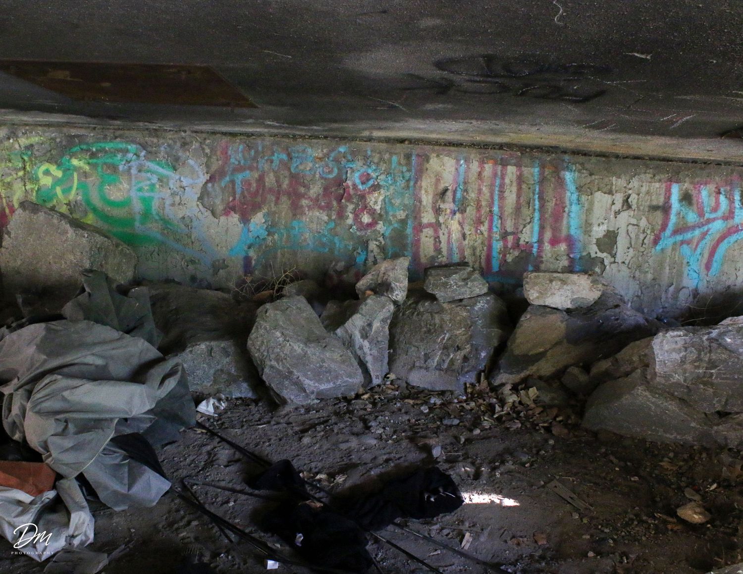 Graffiti covered concrete wall with scattered rocks and debris underneath a bridge.