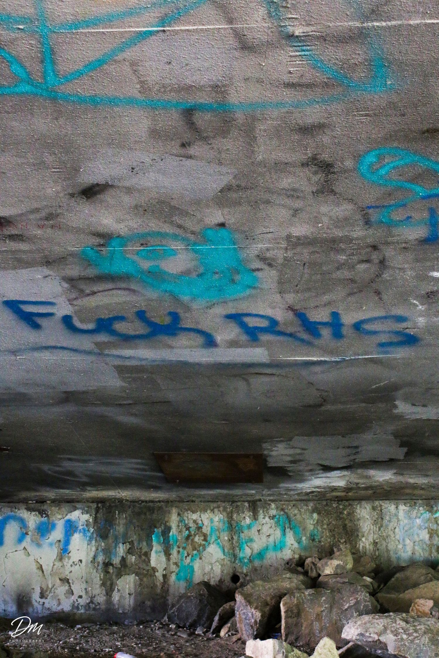 Blue graffiti text on a weathered concrete wall under a bridge near rocks.