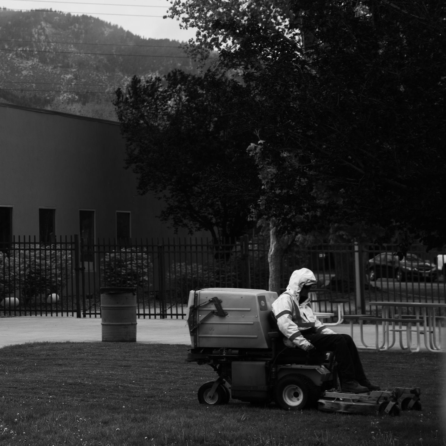 Worker rides lawn mower on green grass with mountains and fence visible in black and white photo.