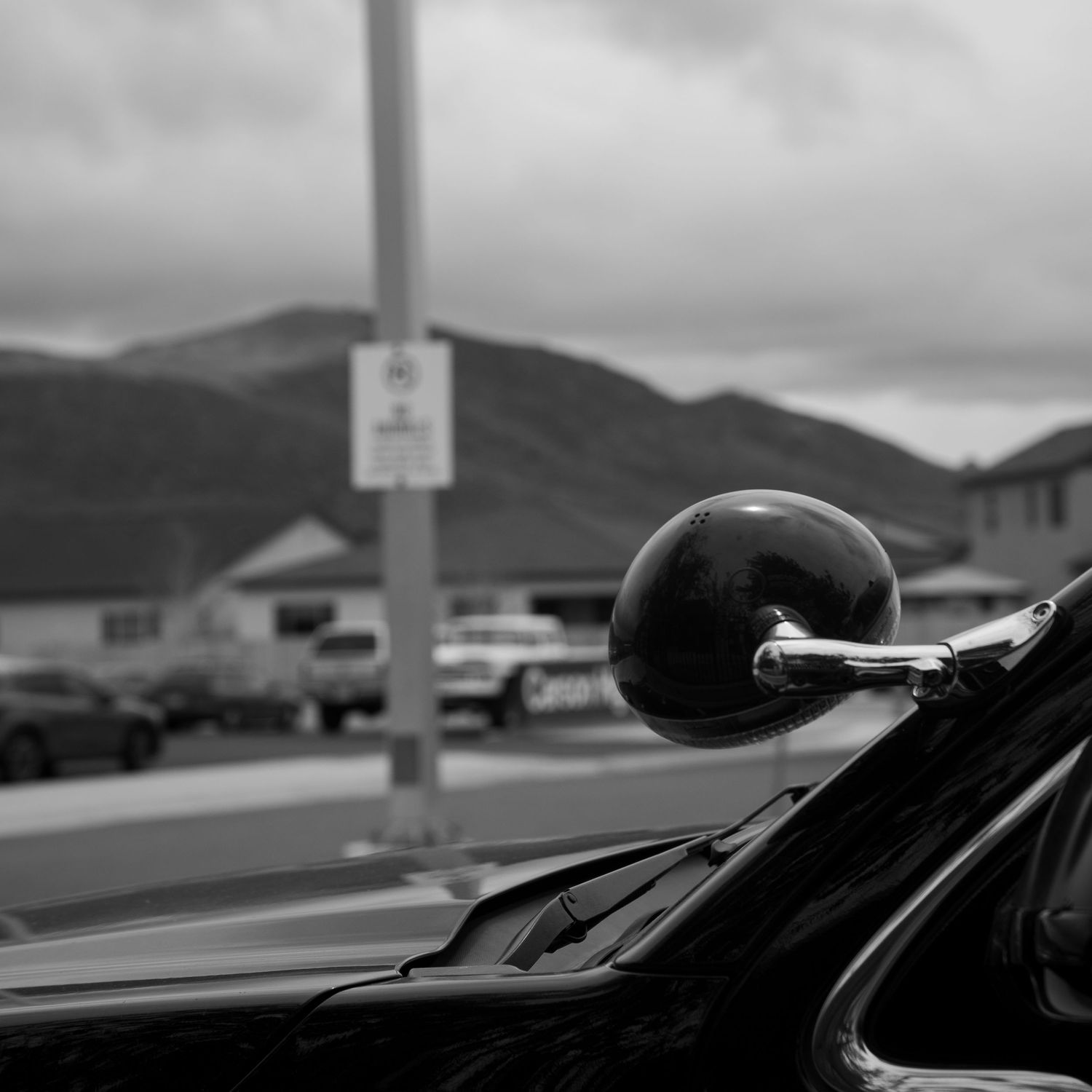 Vintage car side mirror reflects mountains and cloudy sky in black and white street scene.