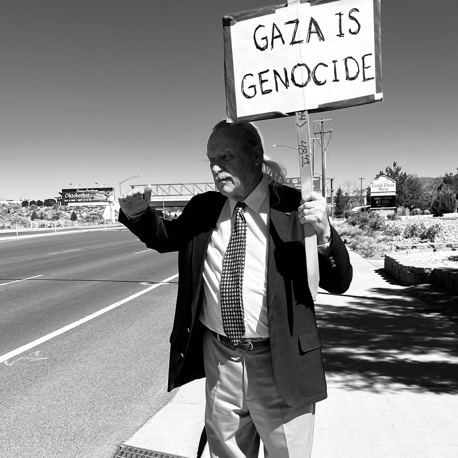 Black and white photo of a man holding a protest sign reading Gaza Is Genocide on a roadside.