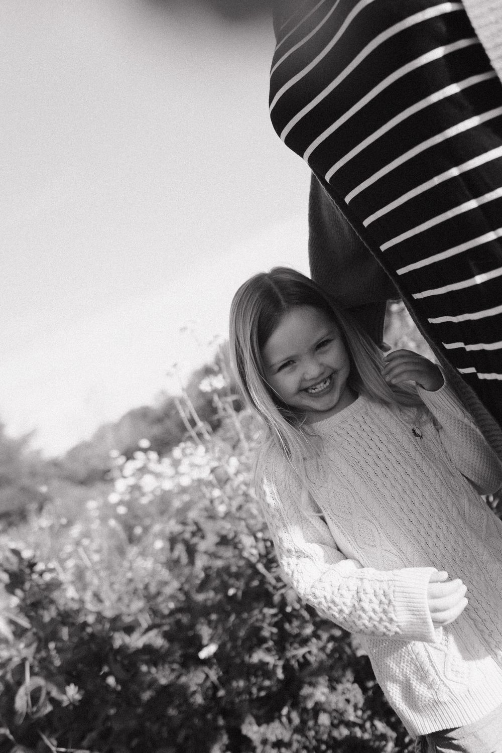 Black and white photo of a child in a white knit sweater smiling and playing outdoors by flowering bushes.