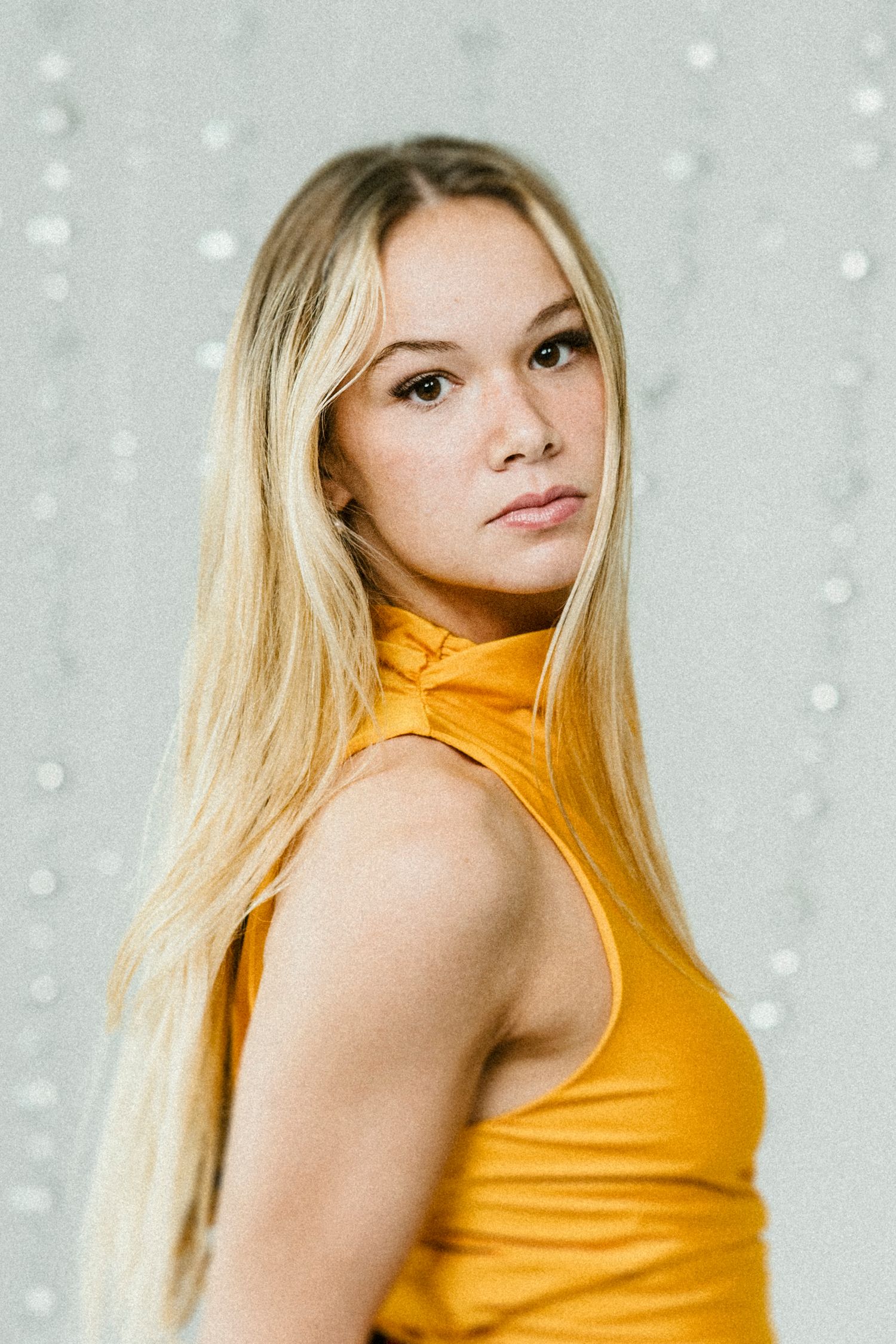 Portrait of a woman with long blonde hair wearing a sleeveless yellow turtleneck against a light textured background with bokeh.