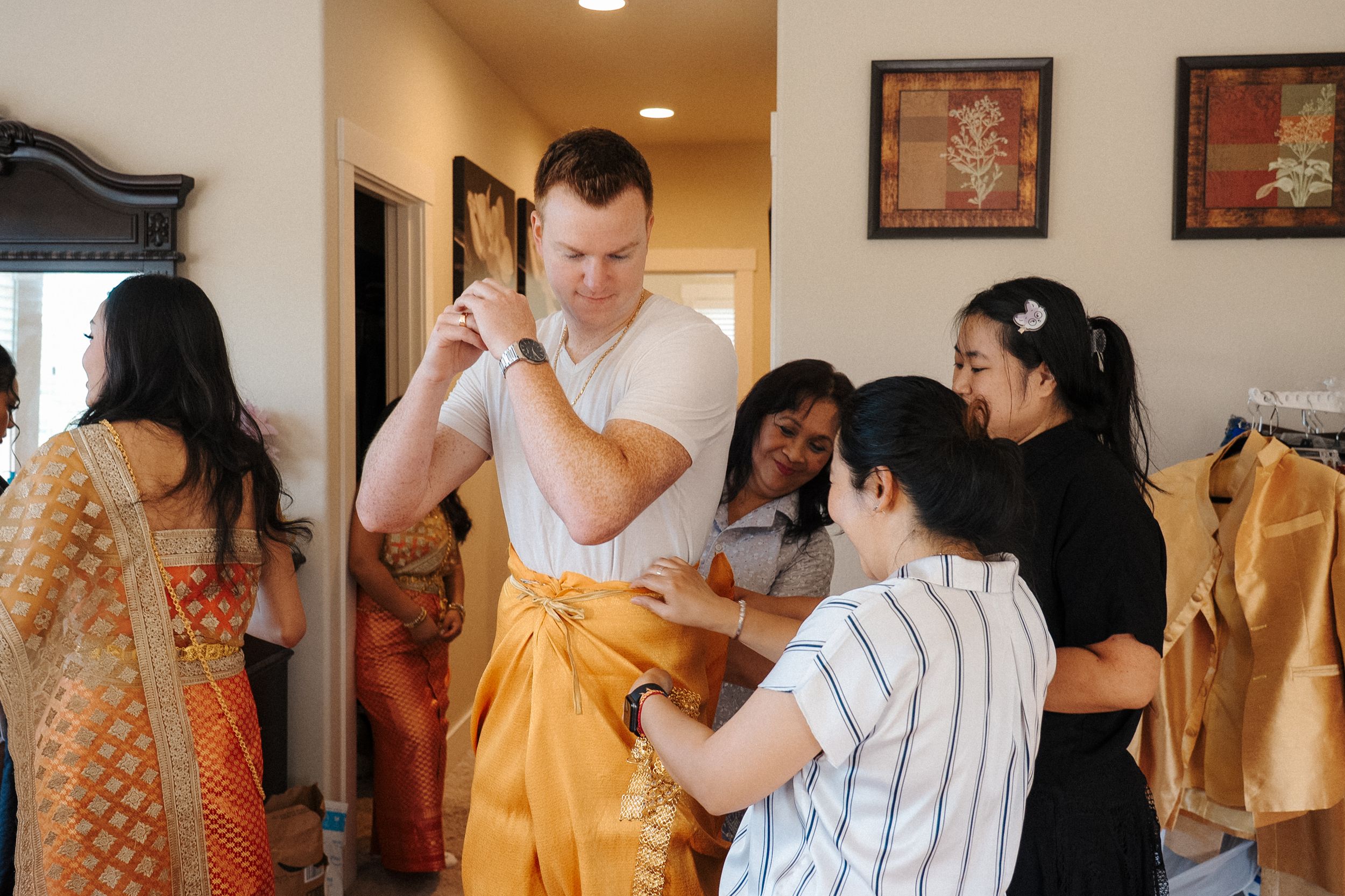 Multiple people help someone get dressed in a traditional gold garment during a formal preparation ceremony.