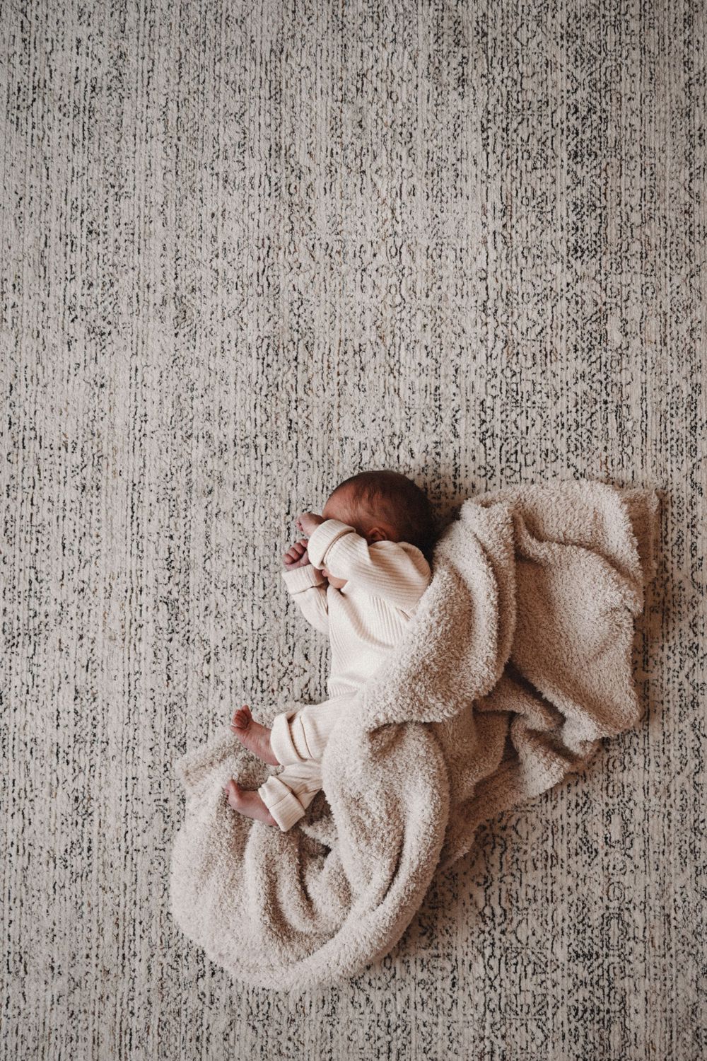 Overhead view of a baby wrapped in soft beige blanket on textured gray carpet.
