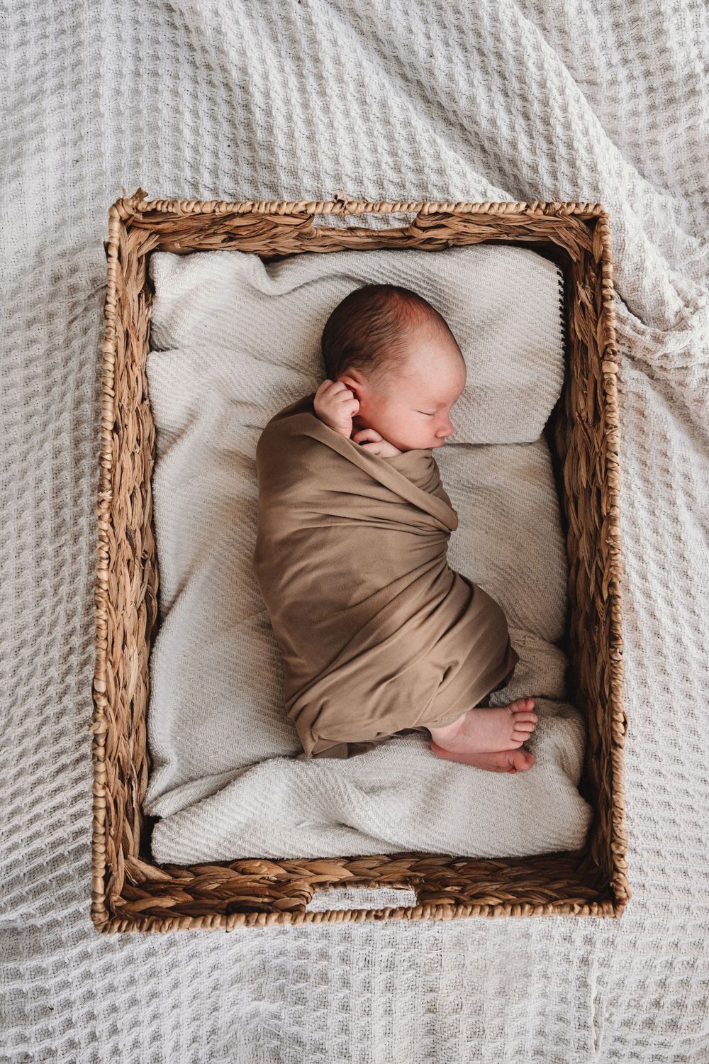 Sleeping newborn wrapped in brown fabric nestled in wicker basket on textured bedding.