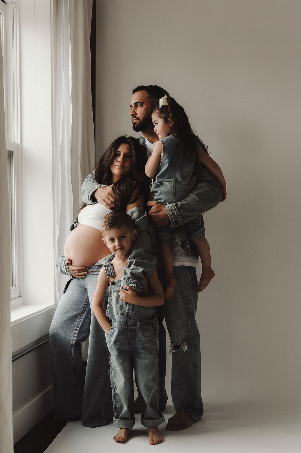 Family in matching denim outfits poses near window for intimate indoor photoshoot.