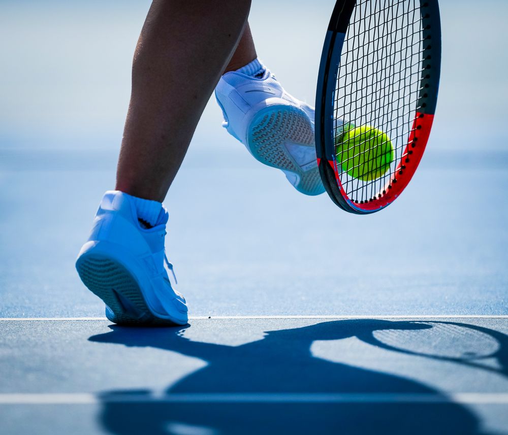 Tennis player in blue shoes prepares to hit a tennis ball with racquet on outdoor court during sunny day.