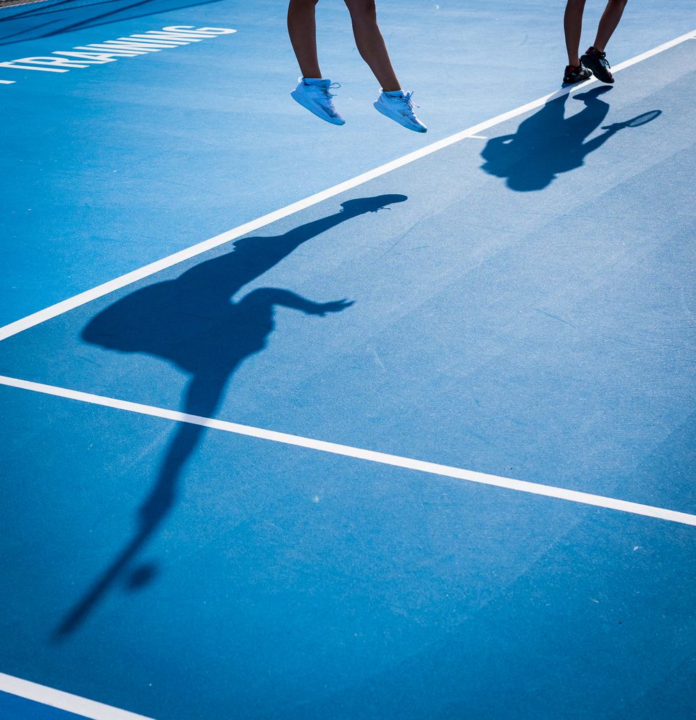 Two people jumping on a blue tennis court creating dramatic shadows during a sunny day.