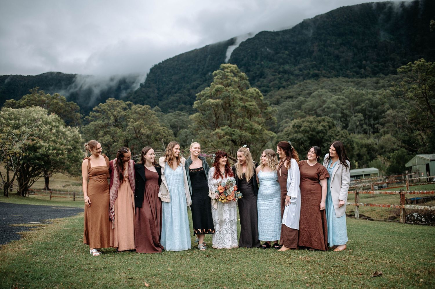 A wedding party stands together outdoors with mountains and lush greenery in the background on an overcast day.