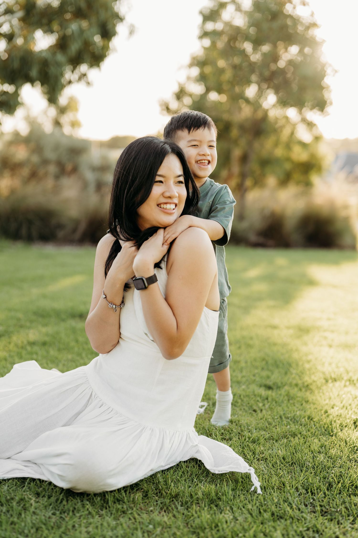 Someone in a white dress lays on grass during sunset while laughing and playing with a small child.