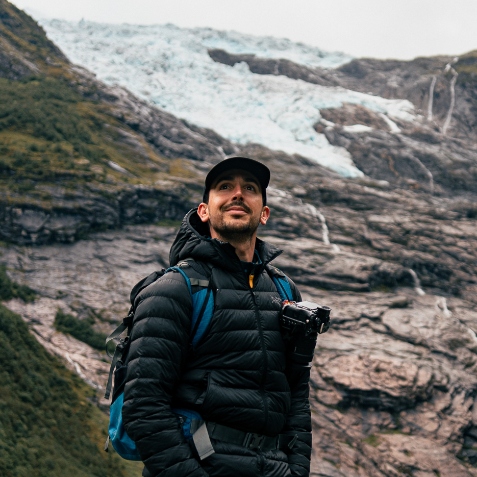 Mountain climber photographer filmmaker wearing black winter coat stands against snowy glacier landscape in sweeping panoramic view.