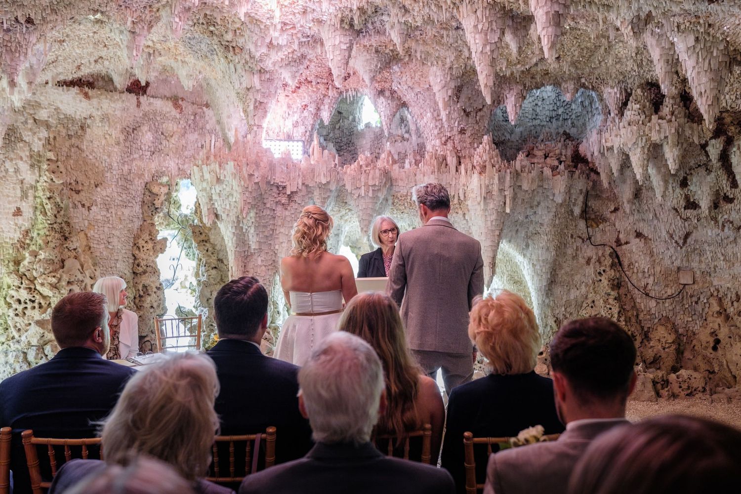 People seated in chairs gather inside an ancient cave-like room with rough stone walls illuminated by pink lighting.