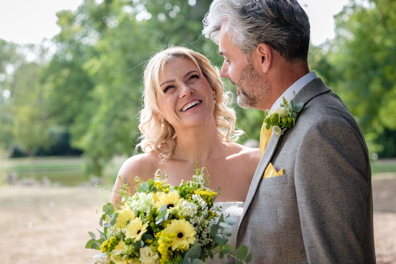 Two people in wedding attire share a romantic moment outdoors with yellow sunflower bouquet and green background.
