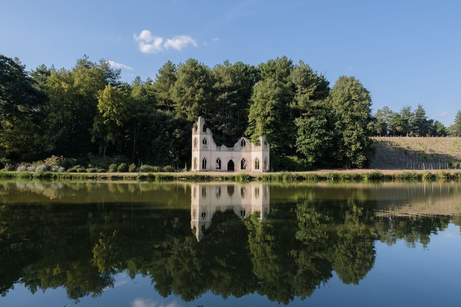 Stone castle tower with arched windows reflects in calm lake waters surrounded by lush green summer trees.