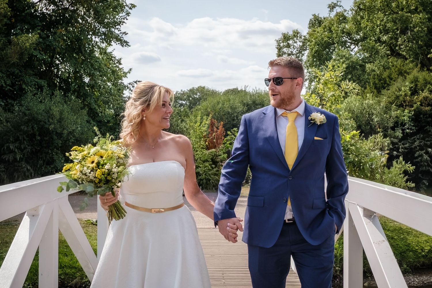 A couple walks hand in hand on a white wooden bridge, with the bride holding yellow flowers and groom in navy suit.