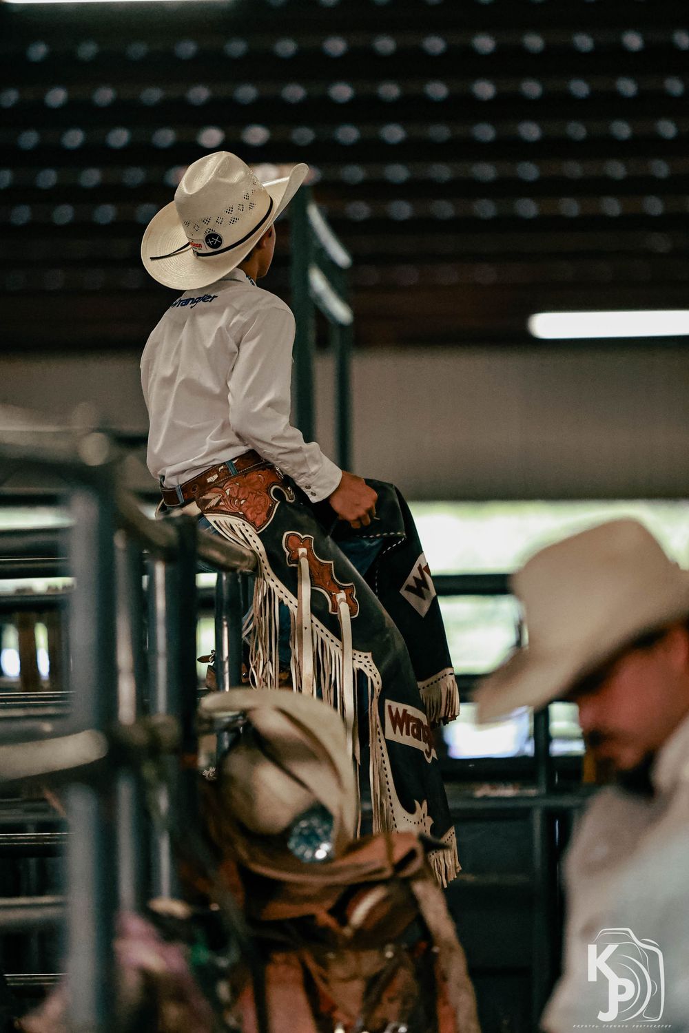Young cowboy in white hat and chaps sits atop a bull at a rodeo event, with another cowboy visible nearby.