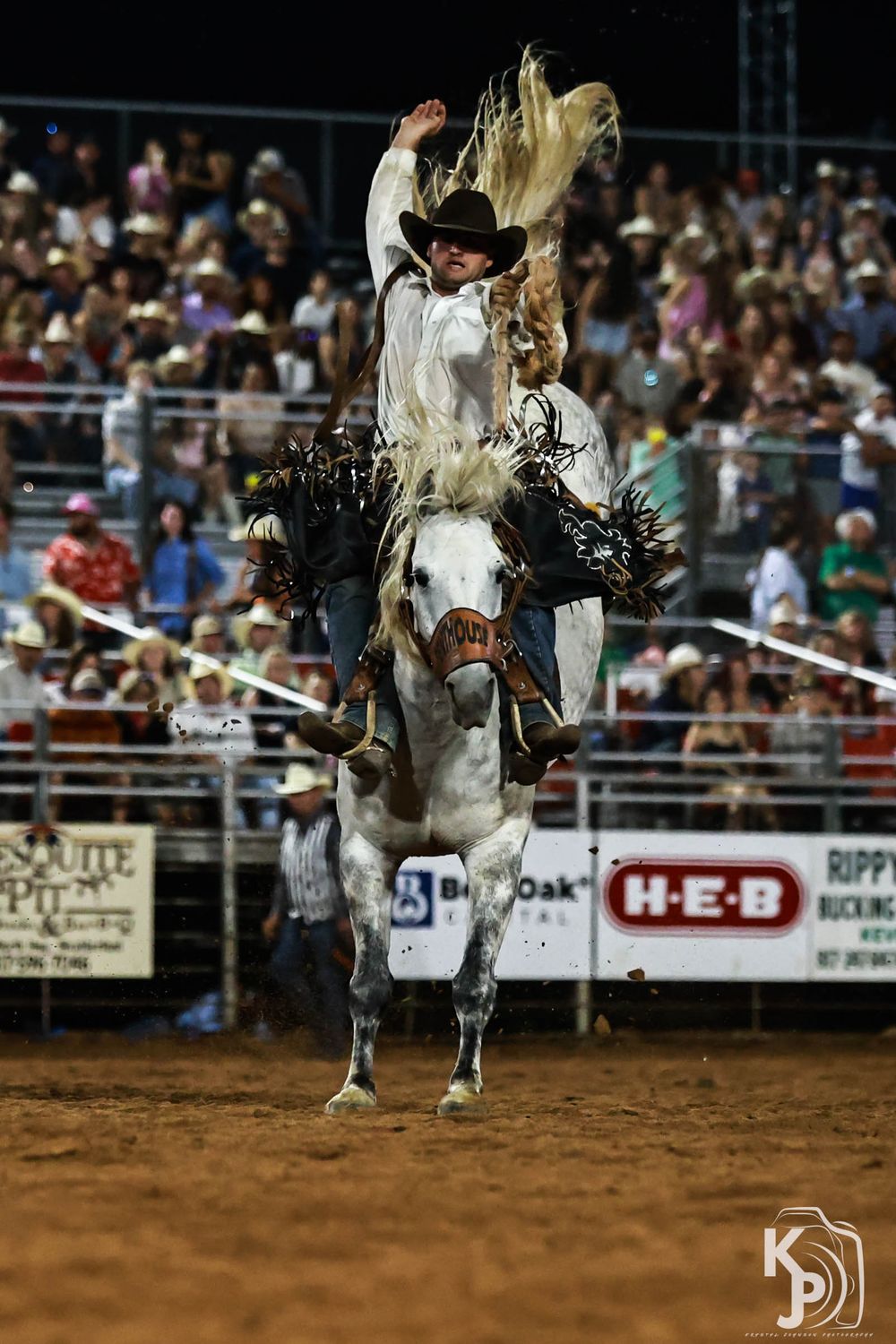 Cowboy riding a bucking horse at a rodeo competition at night, wearing a black hat, with crowd watching in the background.