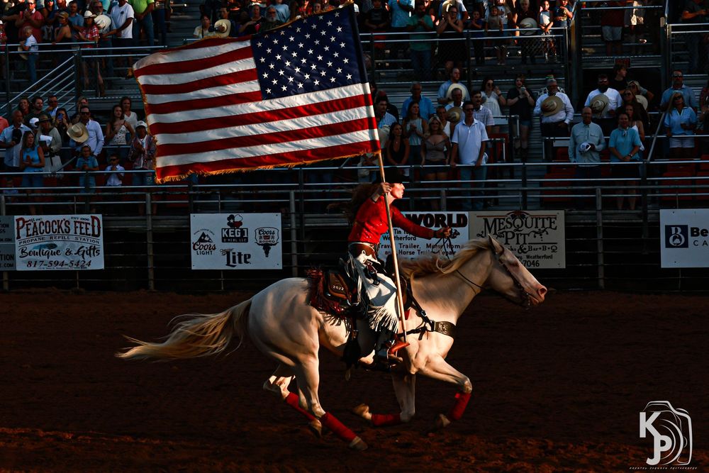 Rider on white horse carrying American flag at rodeo arena with crowd in background.
