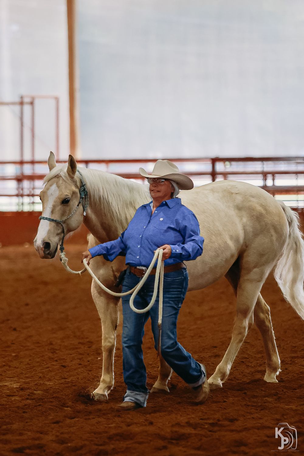 A person in western attire leads a beautiful white horse across a dirt arena during training or practice.