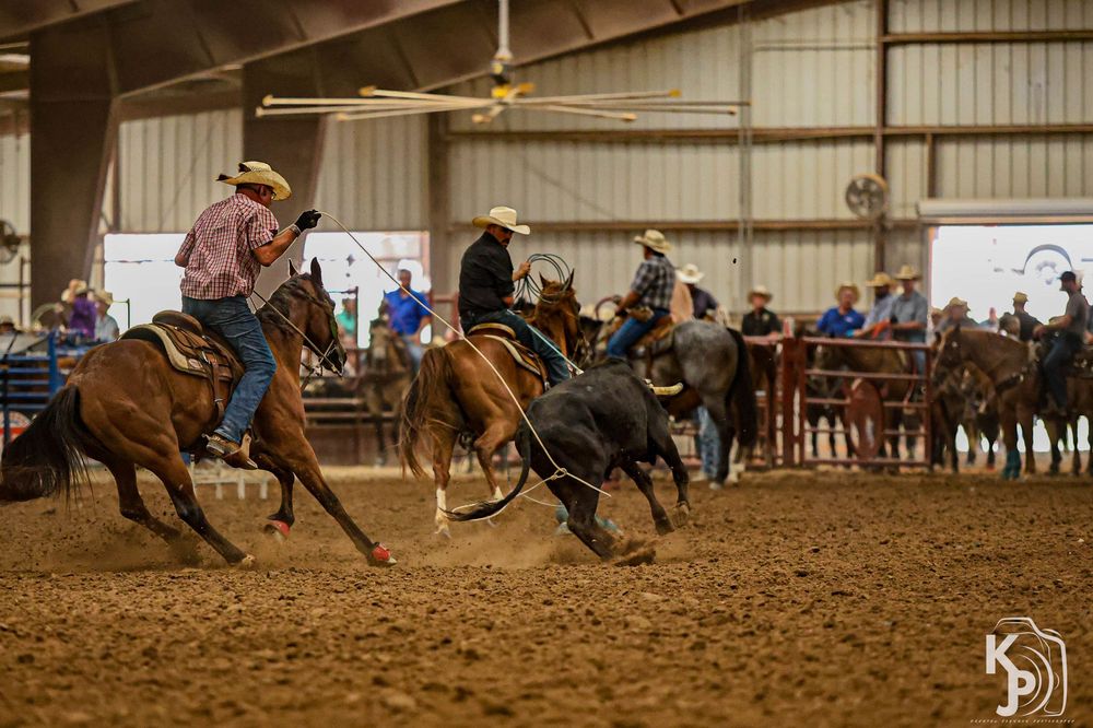 Cowboys on horseback rope a steer during an indoor rodeo competition on a dirt arena floor.