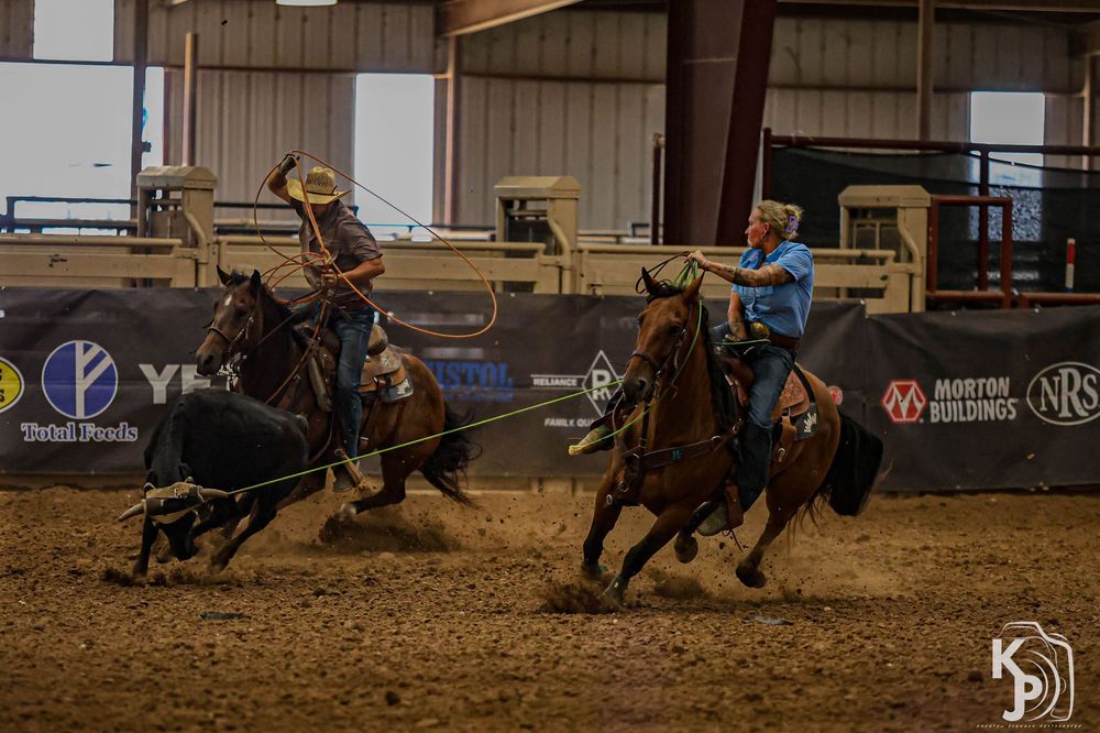 Two cowboys on horseback compete in team roping event inside an indoor rodeo arena.