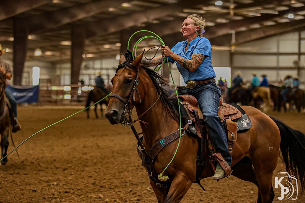 A cowgirl on horseback swings a green lasso rope during an indoor rodeo competition in a sandy arena.