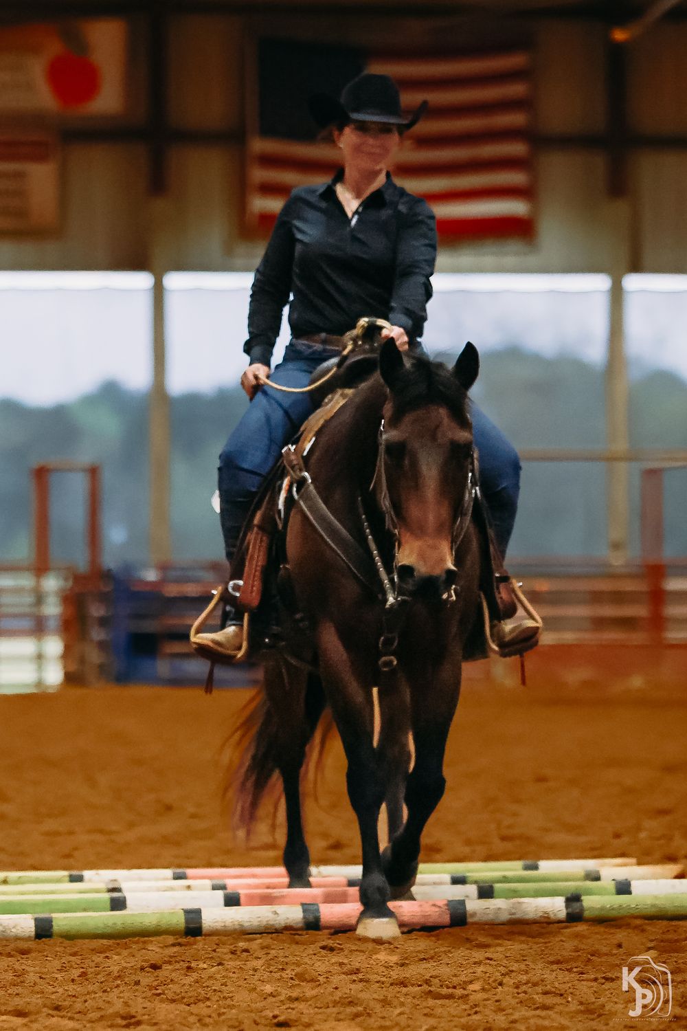 Equestrian riders practice jumping exercises with horses in an indoor arena with American flags on display.