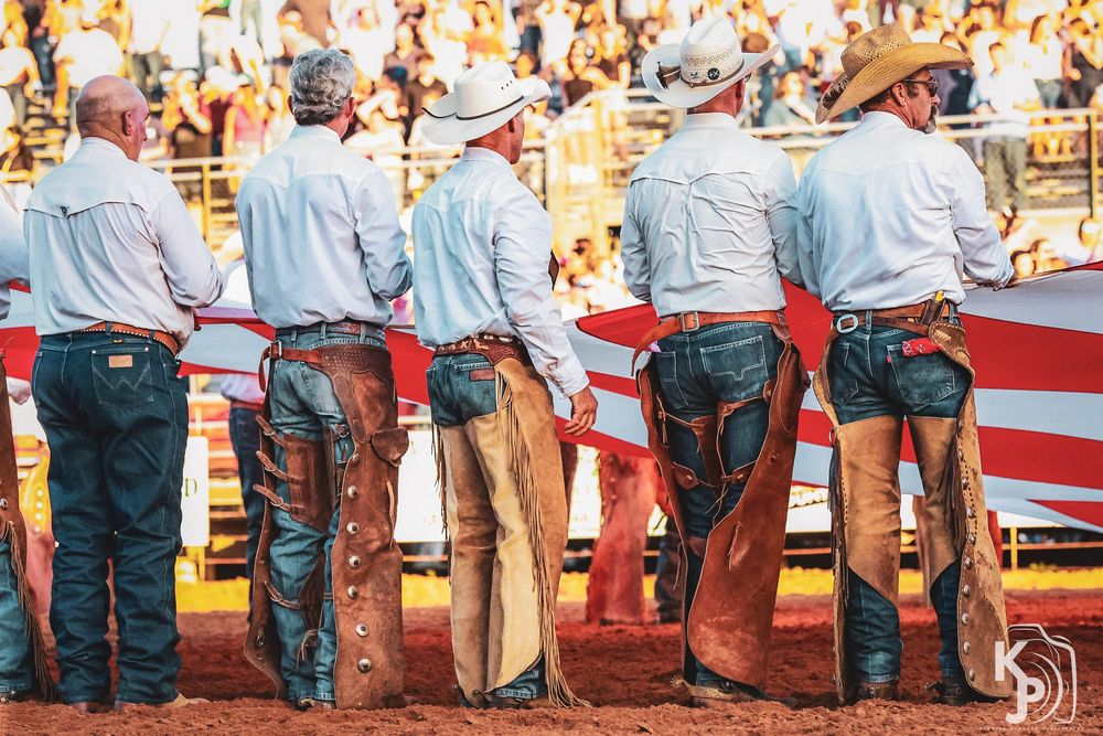 Cowboys in chaps and white shirts hold an American flag at a rodeo arena during a patriotic ceremony.