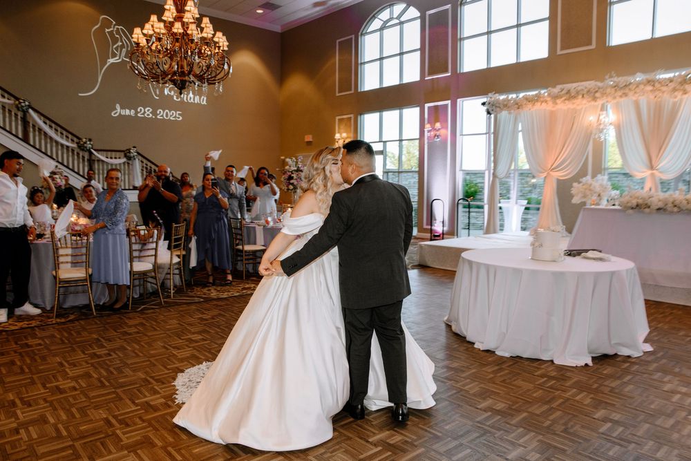 A couple shares their first dance together in an elegant ballroom with a chandelier and arched windows.