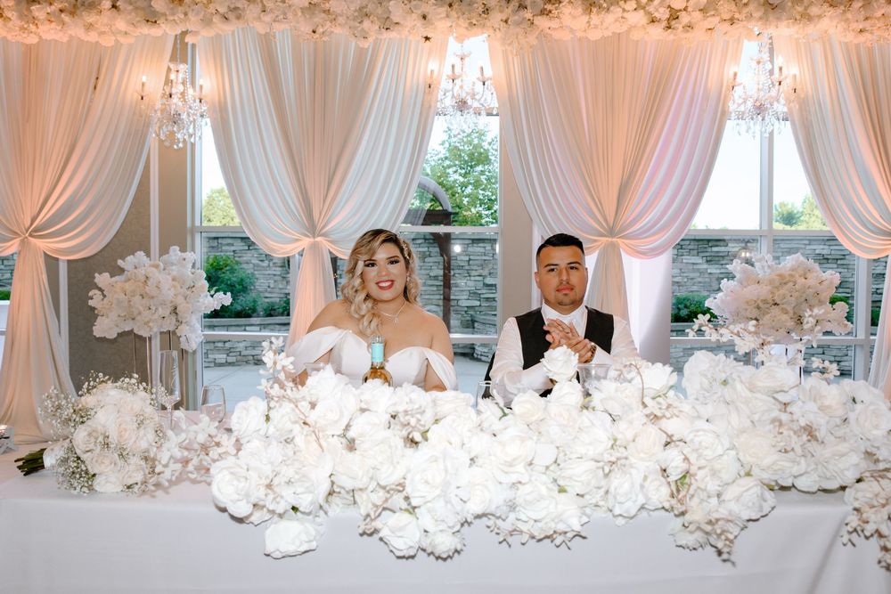 Elegant wedding reception features crystal chandeliers, white floral centerpieces, and sheer drapes creating a romantic atmosphere.