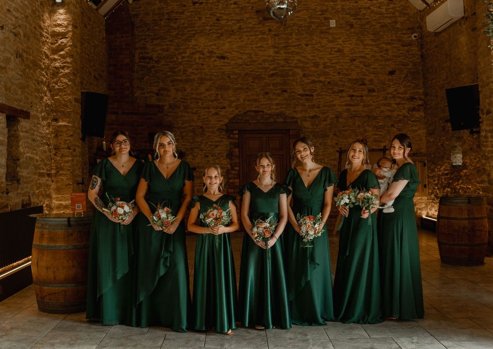 Six bridesmaids in elegant emerald green gowns holding floral bouquets pose together in a rustic stone barn venue.
