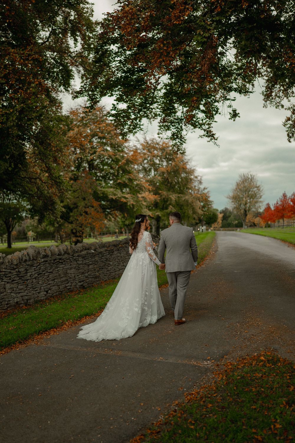 Bride and groom walk hand in hand down a tree-lined path surrounded by autumn foliage on their wedding day.