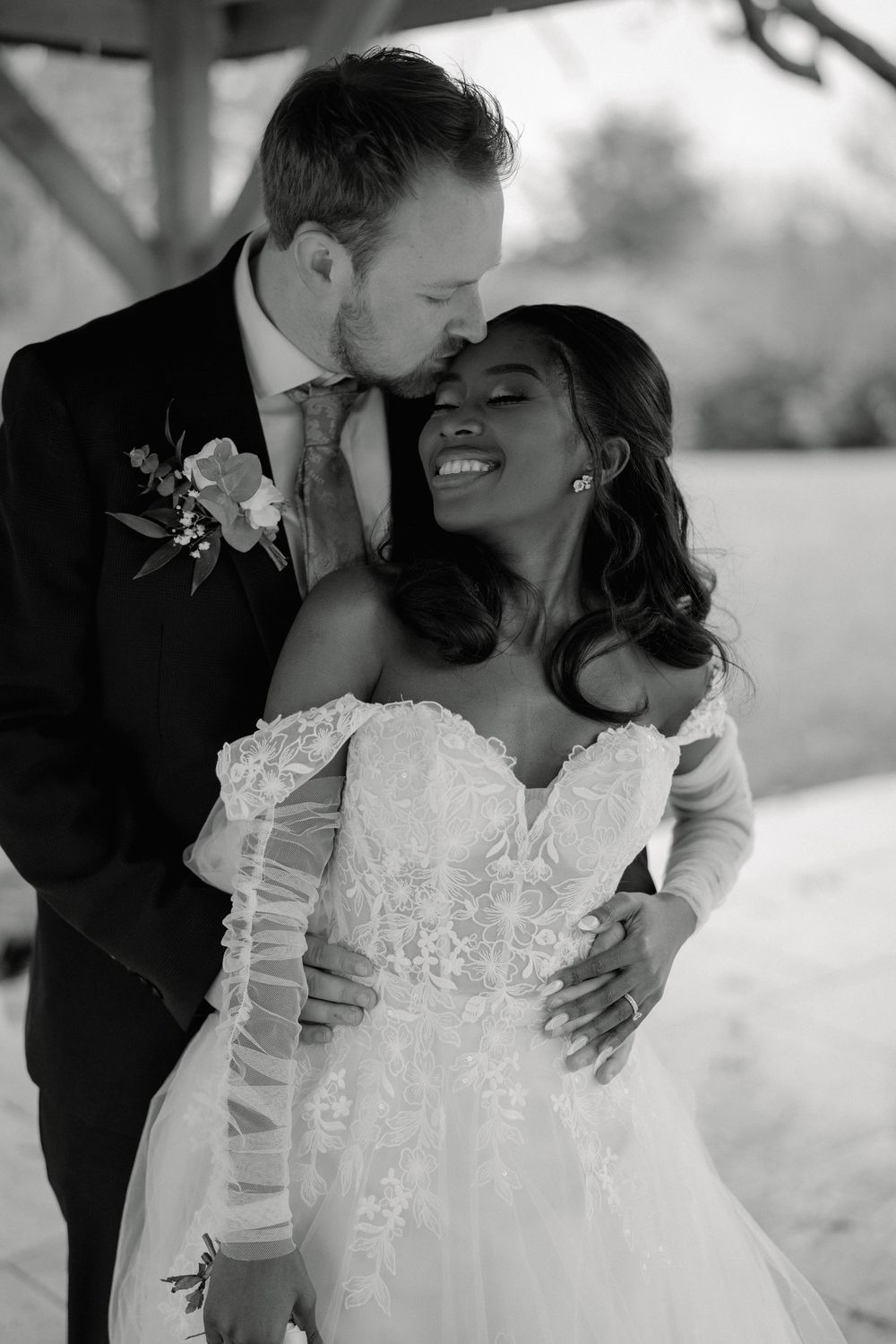 Black and white wedding photo of smiling bride in lace gown and groom in suit sharing a tender moment.