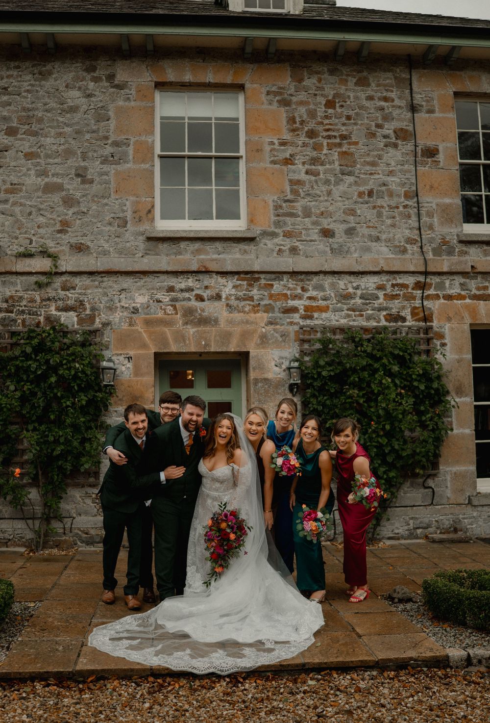 Bride and groom pose with wedding party outside a rustic stone building, bridesmaids in jewel-toned dresses holding bouquets.