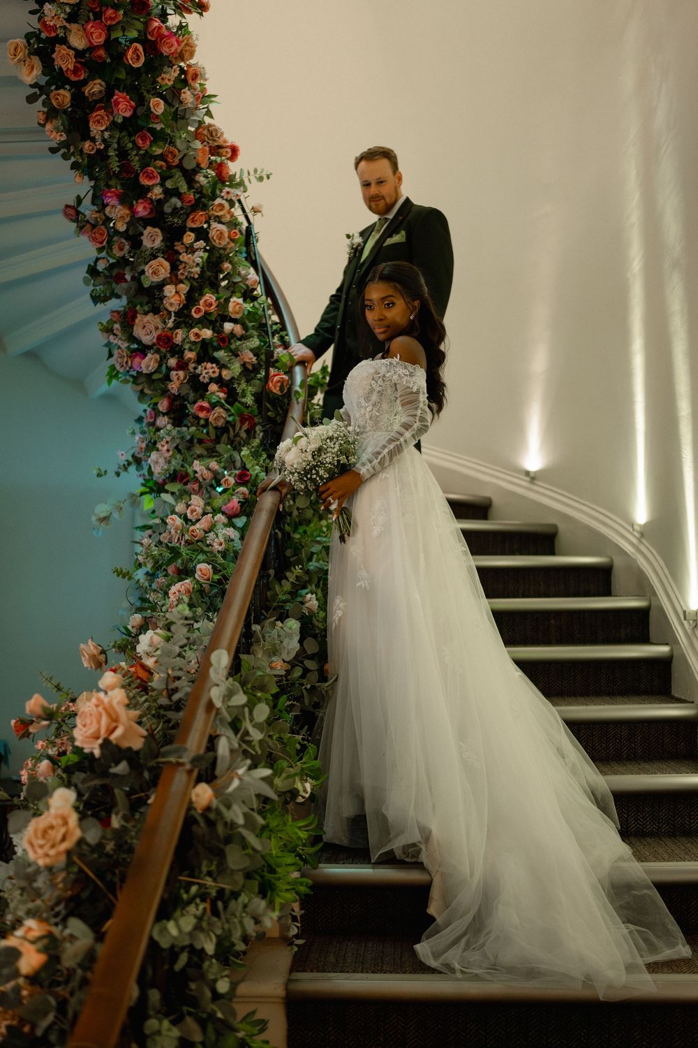 Bride and groom pose on floral-adorned staircase, bride in white tulle gown holding bouquet, groom in dark suit.