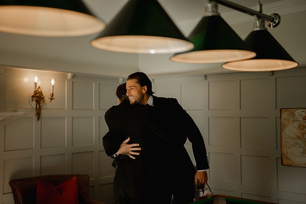 Two people in formal attire share a warm embrace under green pendant lights in an elegant wood-paneled room.