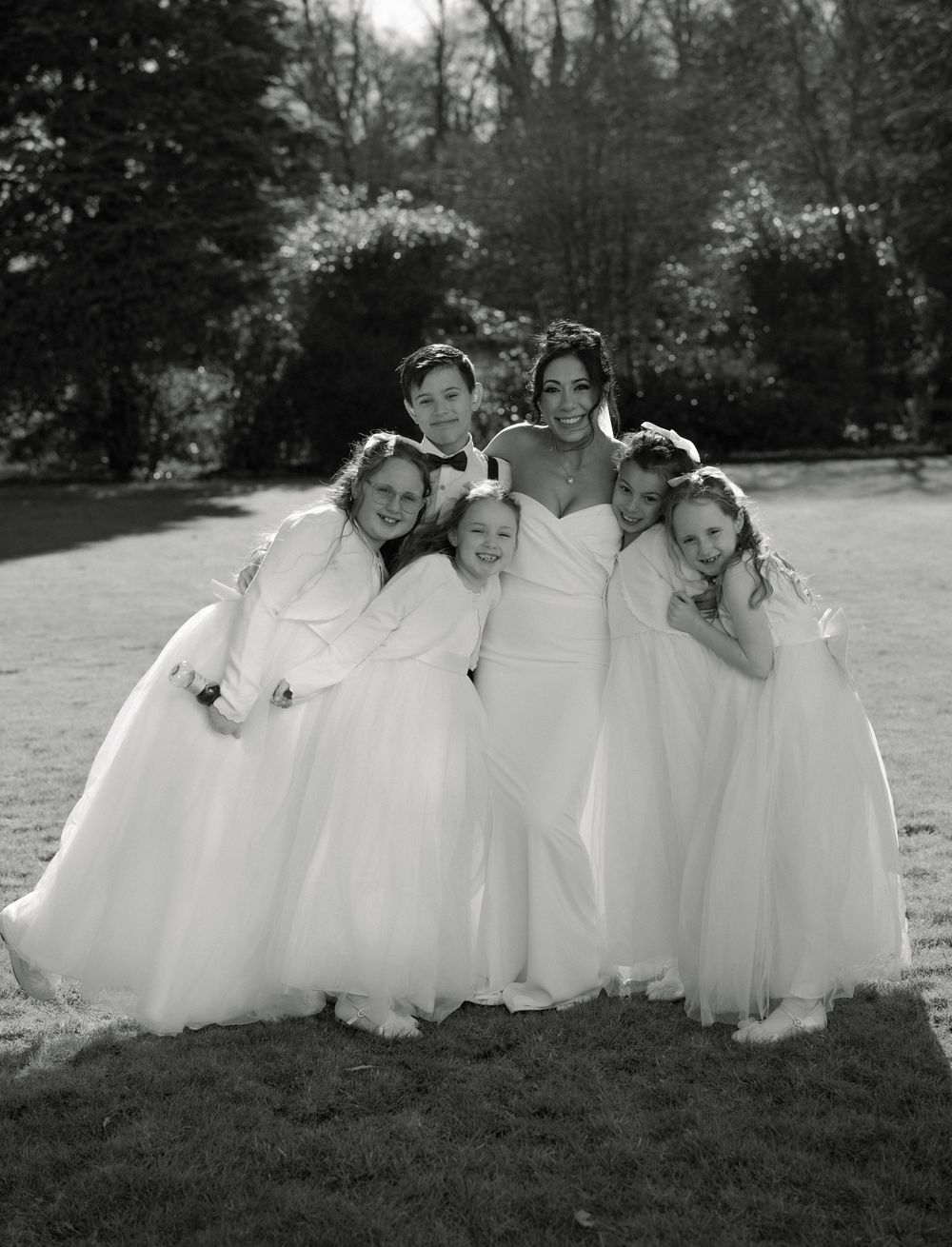 Black and white photo of a bride in a white gown posing with five young flower girls in white dresses outdoors.
